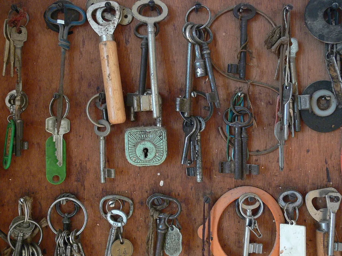 Een verweerd houten bord aan een muur waarop oude sleutels, een hangslot en enkele sleutelringen aan losse haken hangen.