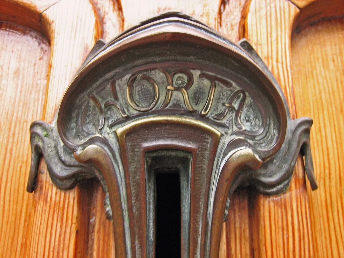 Een sierlijke messing brievenbusklep in een houten voordeur, met de naam Horta in het metaal verwerkt.