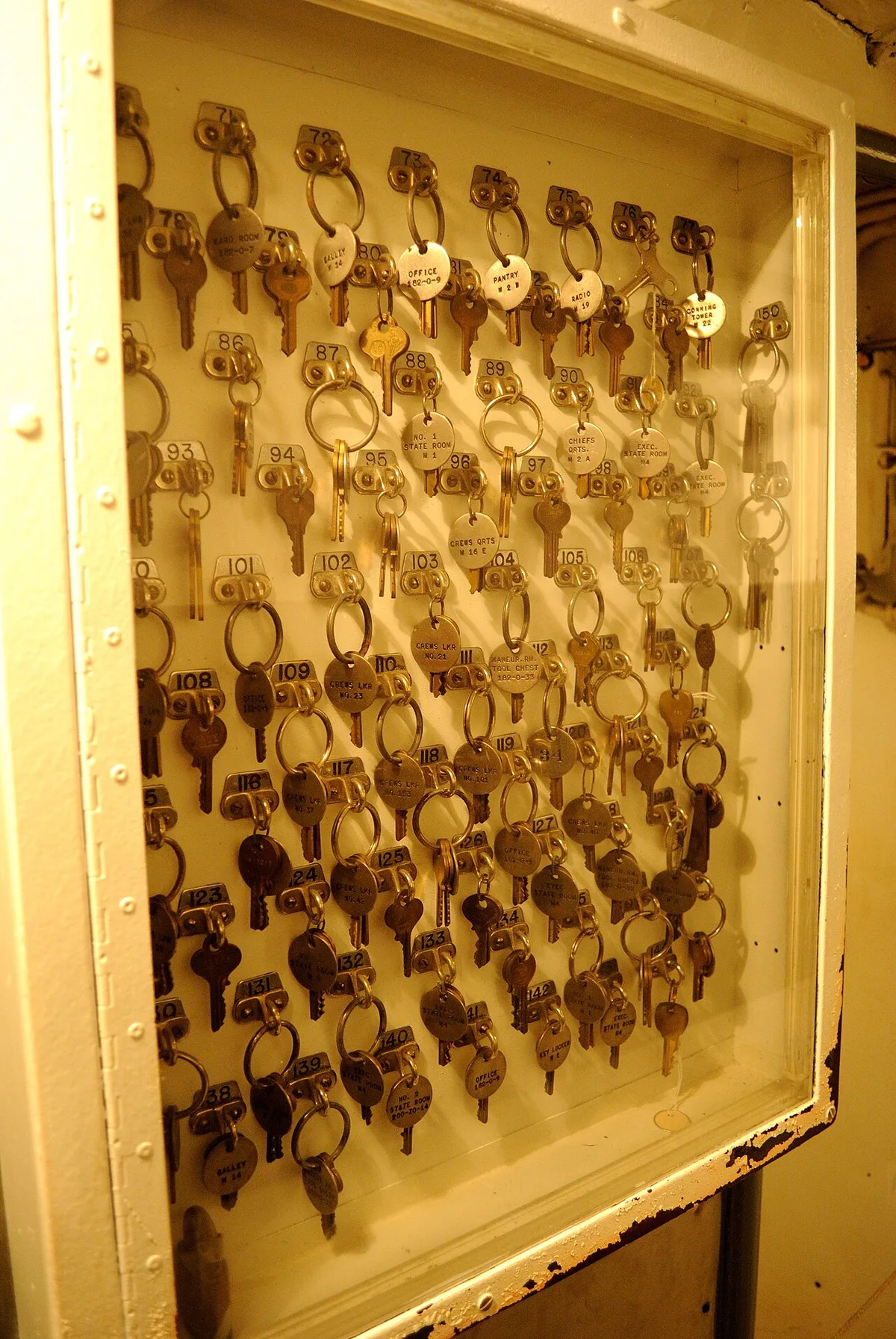 Een geel geschilderd metalen wandpaneel waarop tientallen sleutels aan haakjes hangen, elk met een messing label met een nummer erop.
